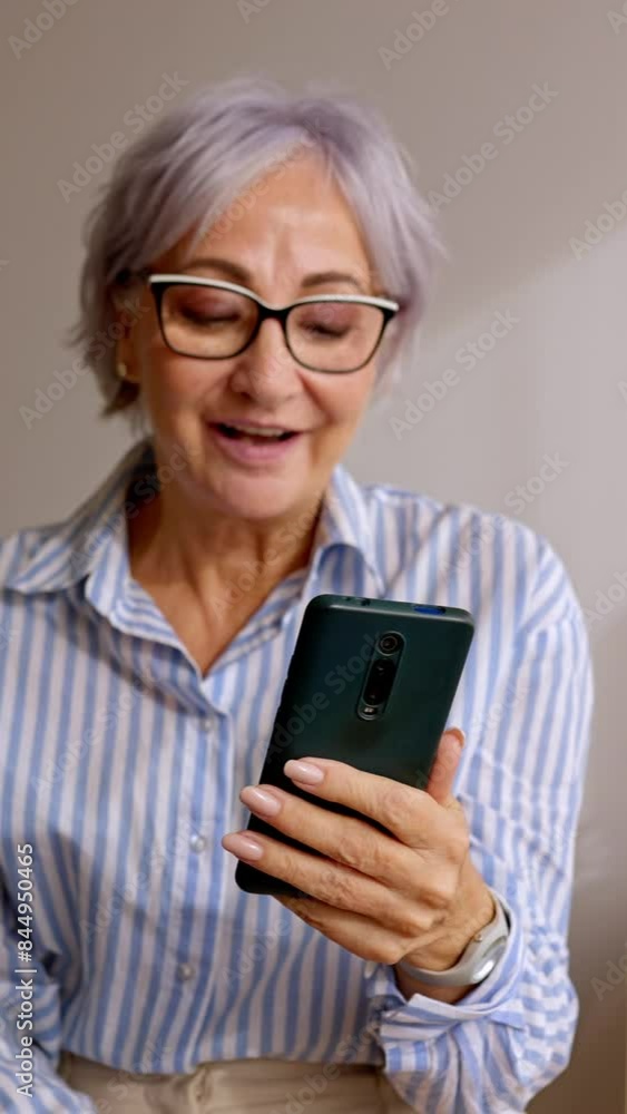 Vertical HD video of happy senior woman using mobile phone while relaxing at home. Smiling cool 60s gray-haired female looking at smartphone screen. Technology and elderly people concept. Vertical HD video of happy senior woman using mobile phone while relaxing at home. Smiling cool 60s gray-haired female looking at smartphone screen. Technology and elderly people concept.