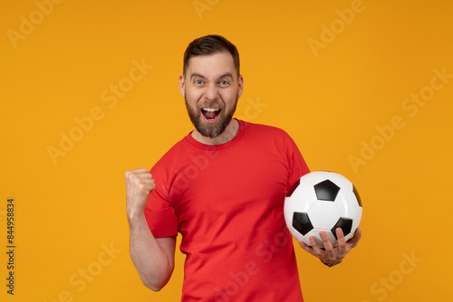 Portrait of euphoric happy football fan man celebrating victory after betting at bookmaker's website, making winner's gesture clenching his fist while holding ball in hand, isolated on yellow backgrou © wpadington