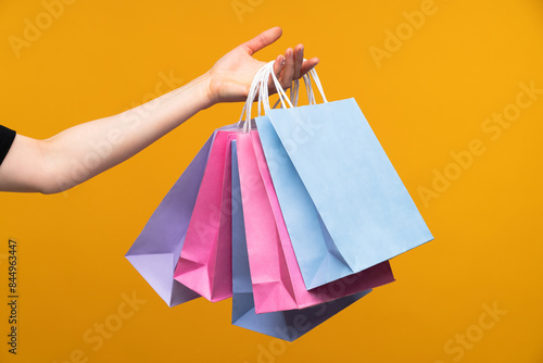 Studio image of female hand holding a bunch of pastel colored papaer shopping bags isolated over bright colored orange yellow background