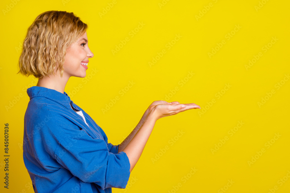 Profile portrait of pretty young lady arms hold empty space wear blue shirt isolated on yellow color background Profile portrait of pretty young lady arms hold empty space wear blue shirt isolated on yellow color background