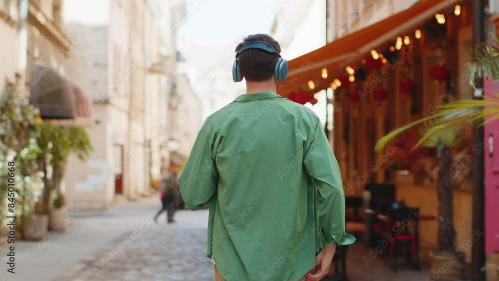 Rear view of happy relaxed man tourist in wireless headphones choosing, listening favorite energetic disco music in smartphone outdoors. Caucasian tourist walking passes by urban sunny city street. Rear view of happy relaxed man tourist in wireless headphones choosing, listening favorite energetic disco music in smartphone outdoors. Caucasian tourist walking passes by urban sunny city street.
