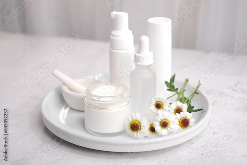 A collection of skincare products on a white tray, including a jar of cream, two pump bottles, and a smaller bottle, all in white packaging. Three daisy-like flowers add a touch of natural beauty.