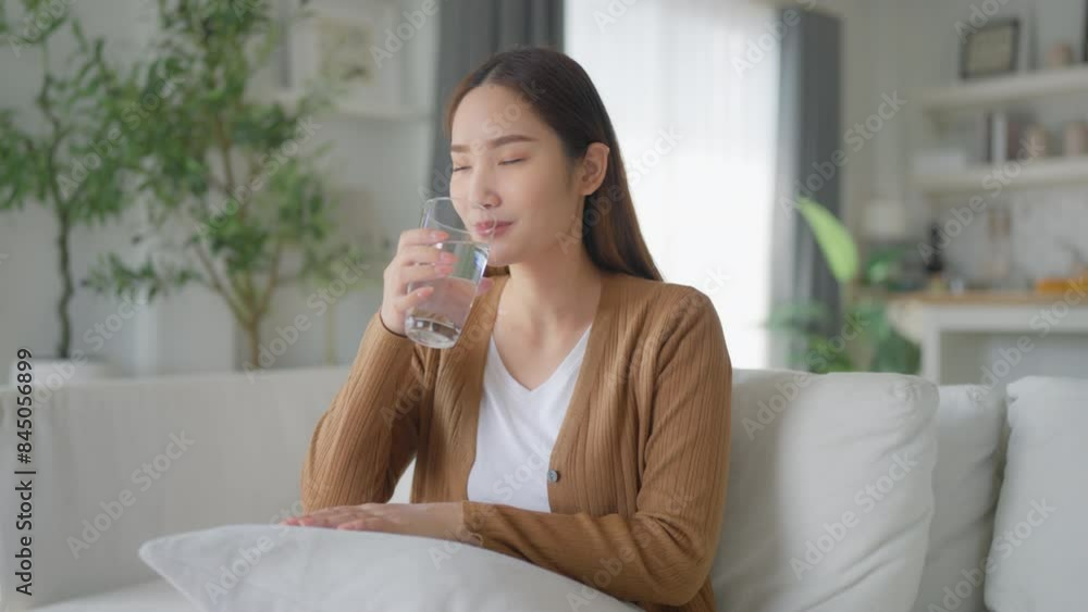 Young Asian woman smiling while sitting on sofa happy expression, hand holding water glass and drinking water Young Asian woman smiling while sitting on sofa happy expression, hand holding water glass and drinking water