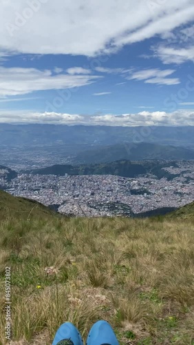 Ascending panoramic view of the city of Quito- Ecuador from Cruz Loma, with the feet of a person sitting on the grass in the foreground video high resolution 4k 60fps