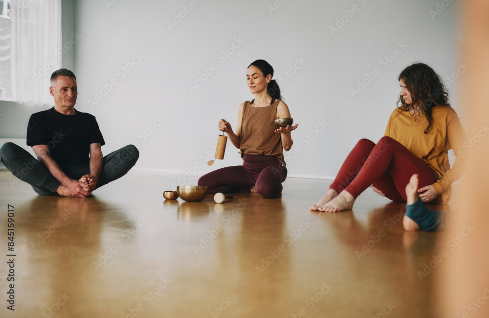 Female yoga instructor using a singing bowl and chime to begin a meditation class of students sitting together on the floor of her studio