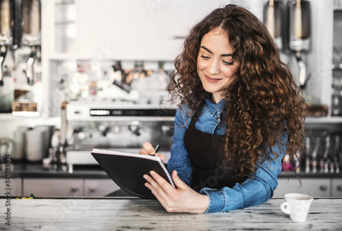 Beautiful female barista studying during part-time work in coffee shop. University student reading school notes while cafe is empty.