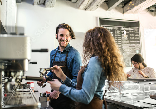 Two young baristas working in coffee shop  standing by counter  preparing coffee.. University students working part-time in cafe.