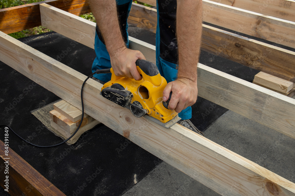 A close-up shot of a carpenters hands using a planer to smooth a wooden beam. The carpenter is wearing blue pants and is focused on their task. A close-up shot of a carpenters hands using a planer to smooth a wooden beam. The carpenter is wearing blue pants and is focused on their task.