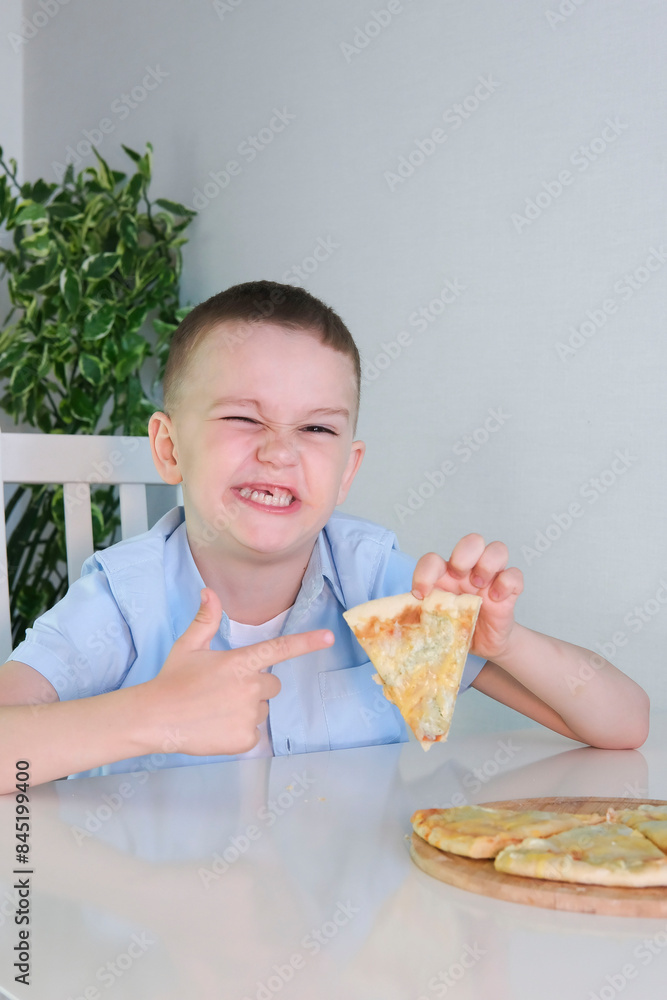 Funny, charming boy refuses to eat pizza. An upset child with facial expression that he doesnt like sits in kitchen at table without an appetite and doesnt want to eat tasteless food. Vertical image. Funny, charming boy refuses to eat pizza. An upset child with facial expression that he doesnt like sits in kitchen at table without an appetite and doesnt want to eat tasteless food. Vertical image.
