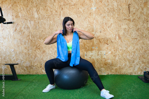 Young and beautiful brunette woman resting while sitting on a gym ball. She has a towel around her neck and is dressed in a top and leggings. Concept of health and sport. photo