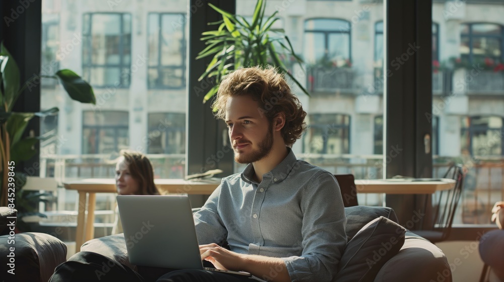 A young man with curly hair and a beard is working on a laptop computer while sitting on a couch in an office. He is wearing a light blue shirt and has a focused expression. The image is taken from a A young man with curly hair and a beard is working on a laptop computer while sitting on a couch in an office. He is wearing a light blue shirt and has a focused expression. The image is taken from a
