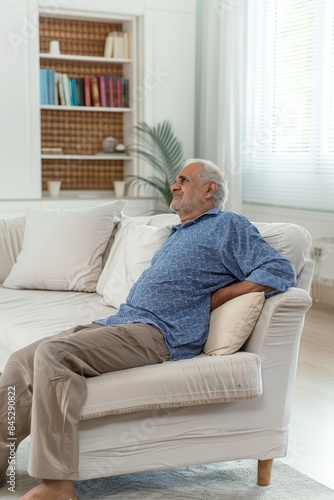 A senior Indian man is sitting in the living room on the sofa with his back pain