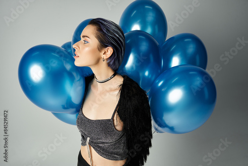 A young woman with short dyed hair balances blue balloons in a stylish studio setting, embodying a whimsical birthday celebration.