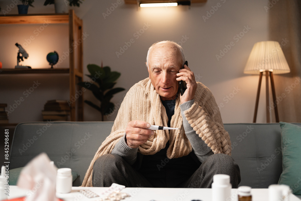 Elderly sick man sitting on couch, checking temperature with thermometer, talking on phone, wrapped in blanket, surrounded by medicine Elderly sick man sitting on couch, checking temperature with thermometer, talking on phone, wrapped in blanket, surrounded by medicine