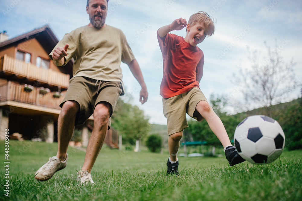 Dad having fun with young son, playing football, running and kicking the ball. Playing on a lawn in front of their house. Fatherhood and Father's Day. Dad having fun with young son, playing football, running and kicking the ball. Playing on a lawn in front of their house. Fatherhood and Father's Day.