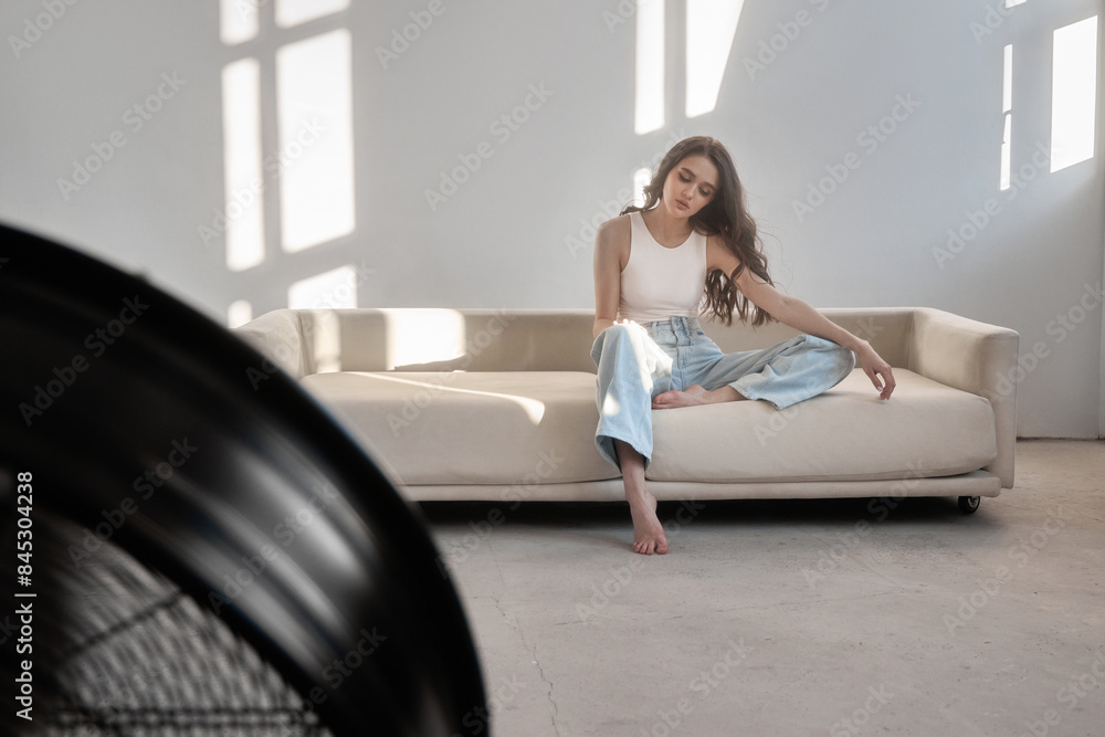 Young fashion woman sits on a couch in a well-lit room. She is wearing blue jeans and a white tank top. The light shines through the window behind her Young fashion woman sits on a couch in a well-lit room. She is wearing blue jeans and a white tank top. The light shines through the window behind her