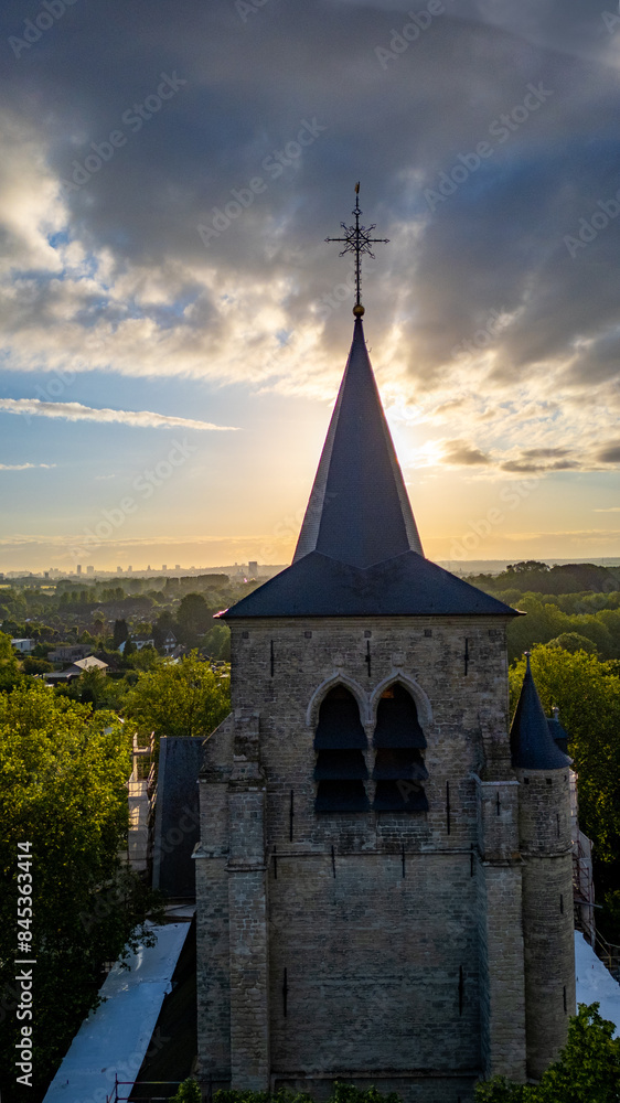 Obraz premium A historic stone church tower stands under a dramatic sunset, surrounded by lush greenery and a serene ambiance