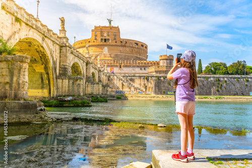 A young girl captures a photo of Castel SantAngelo and a bridge in Rome with her smartphone on a bright sunny day. Rome, Italy