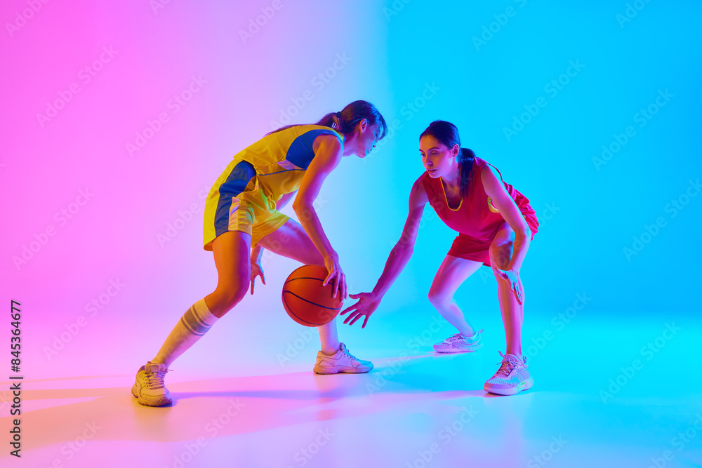 Obraz premium On-Court Tactics. Young women in colorful sportswear, basketball players in motion with ball, competing on gradient pink blue background in neon. Concept of sport, active lifestyle, competition, game
