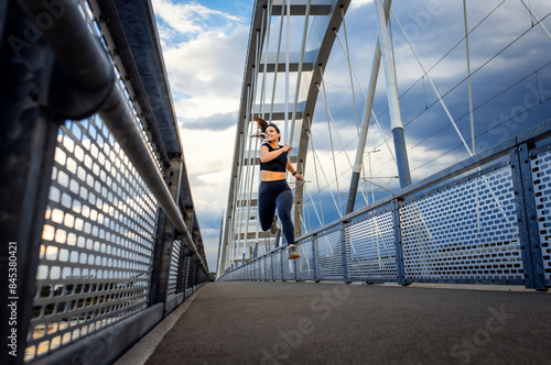 Young woman in black sport outfit running on city bridge.