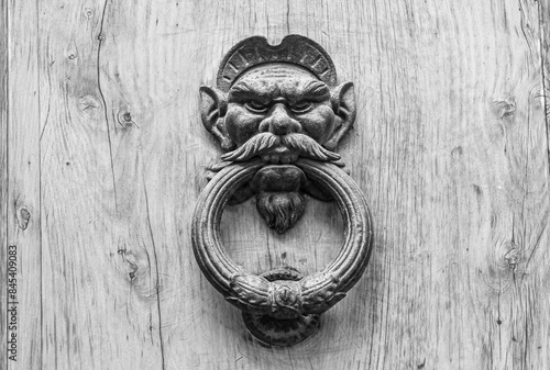 Black and white photo. Close-up of an old wrought iron knocker depicting the head of a satyr or demon on a wooden door, Florence, Tuscany, Italy\