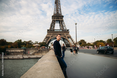 Photography Handsome man in dark pullover is standing on the bridge background of the Eiffel