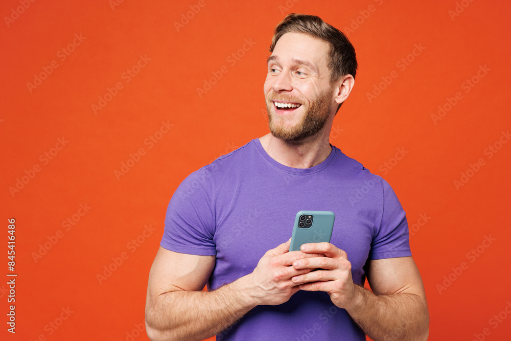 Young fun surprised happy man he wearing purple t-shirt casual clothes hold in hand use mobile cell phone look aside on blank area isolated on red orange background studio portrait. Lifestyle concept. Young fun surprised happy man he wearing purple t-shirt casual clothes hold in hand use mobile cell phone look aside on blank area isolated on red orange background studio portrait. Lifestyle concept.