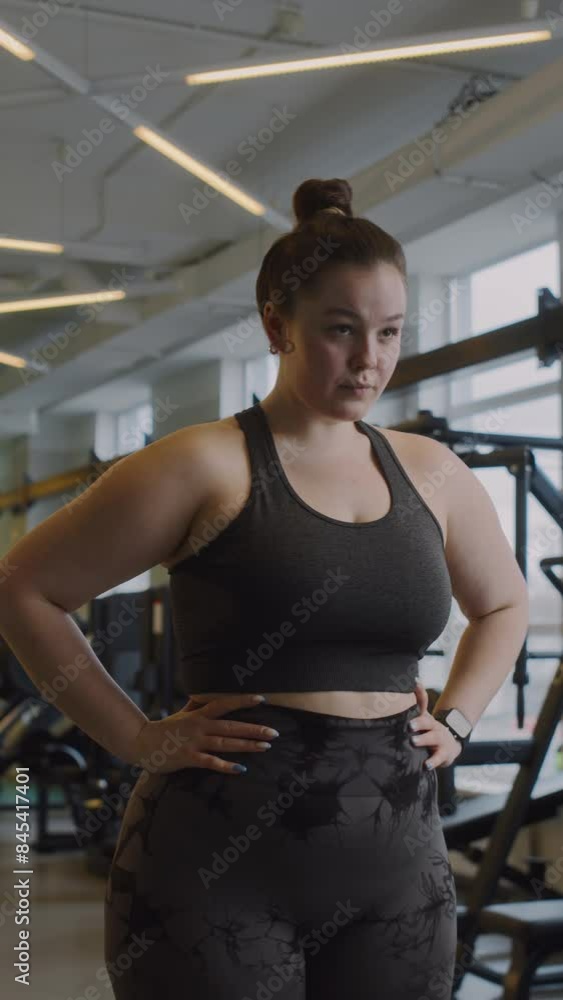 Vertical medium shot of young Caucasian plus size woman in activewear warming up neck and wrists while having gym workout Vertical medium shot of young Caucasian plus size woman in activewear warming up neck and wrists while having gym workout
