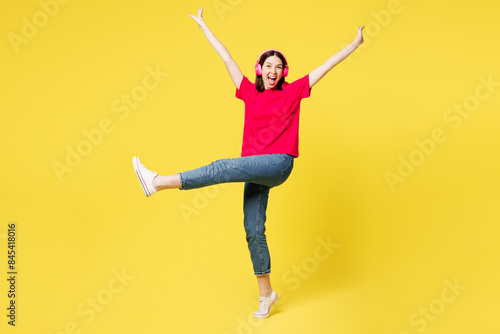 Fotografie Full body young happy woman she wear pink t-shirt casual clothes listen to music in headphones dace raise up legs hands isolated on plain yellow orange background studio portrait