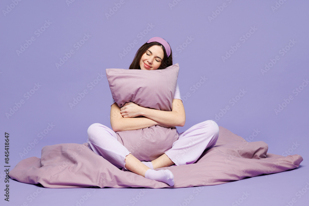 Full body young calm woman wears pyjamas jam sleep eye mask rest relax at home sit on duvet hold hug pillow close eyes isolated on plain pastel purple background studio. Good mood night nap concept. Full body young calm woman wears pyjamas jam sleep eye mask rest relax at home sit on duvet hold hug pillow close eyes isolated on plain pastel purple background studio. Good mood night nap concept.