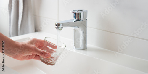 Thirsty woman filling glass with fresh pure drinkable tap water from faucet in white bathroom, banner with concept of natural clean aqua at home