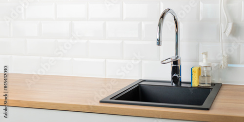 A clean empty black kitchen sink in a kitchen with a chrome faucet, wooden countertop, and white subway tile backsplash with dishwashing liquid, brush and sponge, banner with copy space