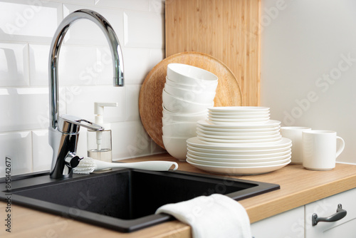 Clean dry washed tableware. Concept of washing dishes. Dishwashing liquid with brush, plates and cups near black kitchen sink with a chrome faucet on wooden counter and white subway tile backsplash