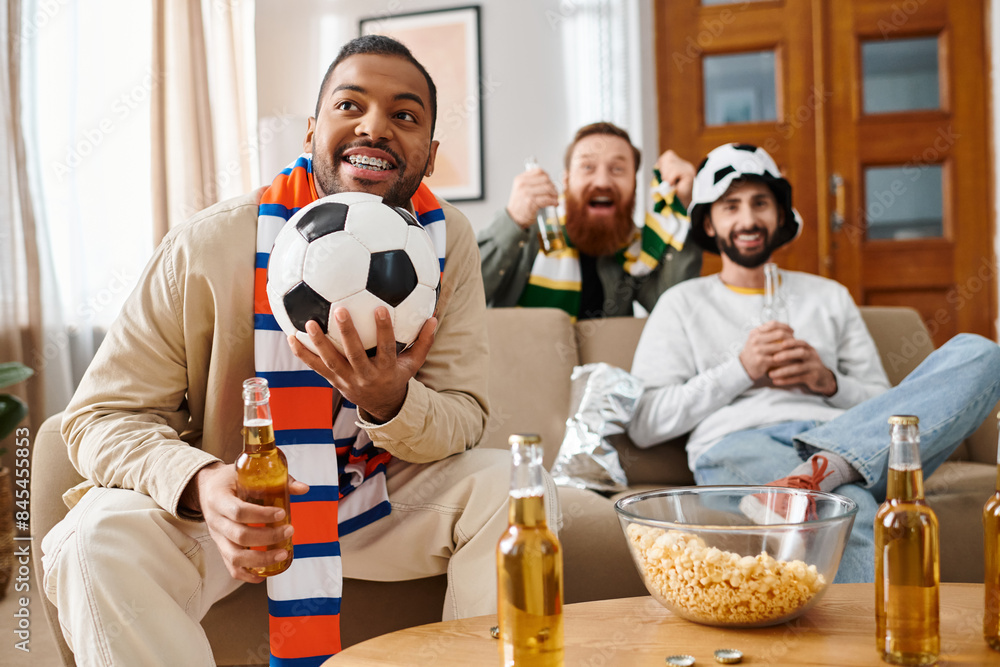 A man in casual attire holds a bottle of beer while holding a soccer ball, enjoying a cheerful moment with friends at home.