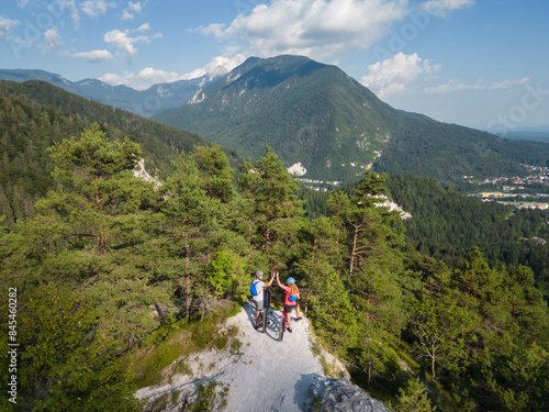 Tablou pe pânză Biker couple after electric mountain bike ride enjoying the spectacular view and success, with a gesture of high five on the hilltop, aerial view