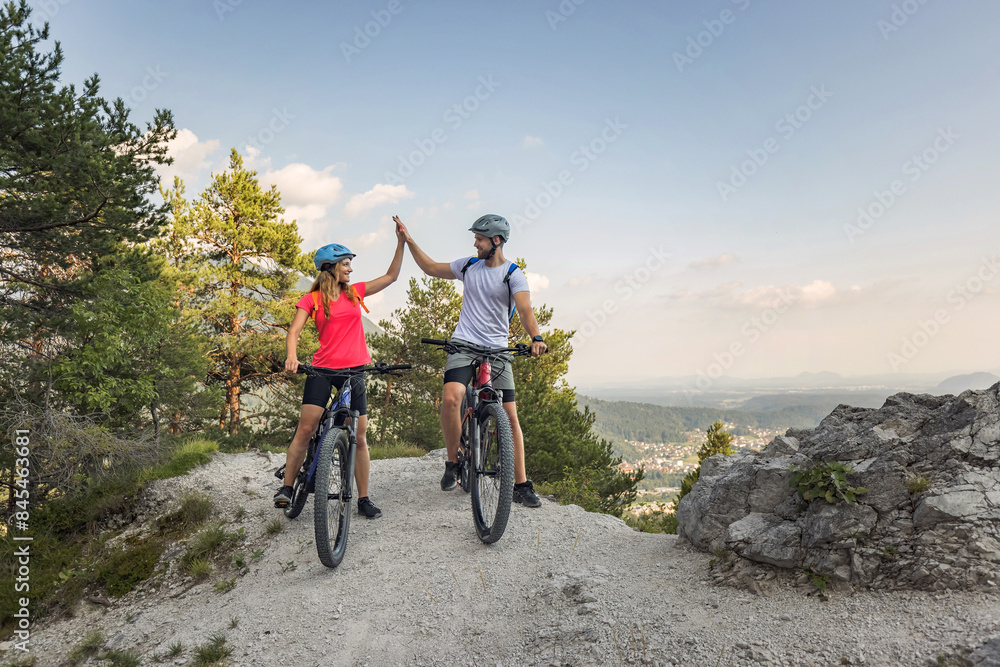 Fototapeta premium Young couple enjoying spectacular scenery, smiling while sitting on electric mountain bikes and giving a high five to each other.