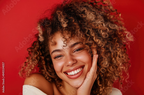 A beautiful woman with curly hair smiles and looks at the camera, against a red background.