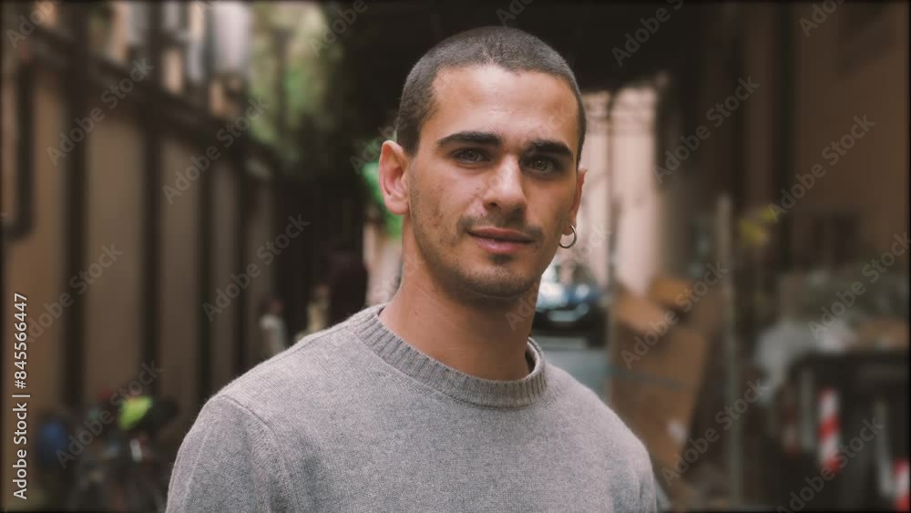 20-year-old man with short hair, wearing a gray sweater, standing in a suburban courtyard, looking at the camera and then smiling. 20-year-old man with short hair, wearing a gray sweater, standing in a suburban courtyard, looking at the camera and then smiling.