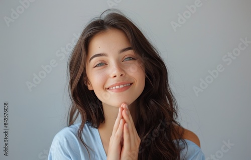 Young Woman With Long Brown Hair Smiles and Holds Hands Together in Front of Grey Wall