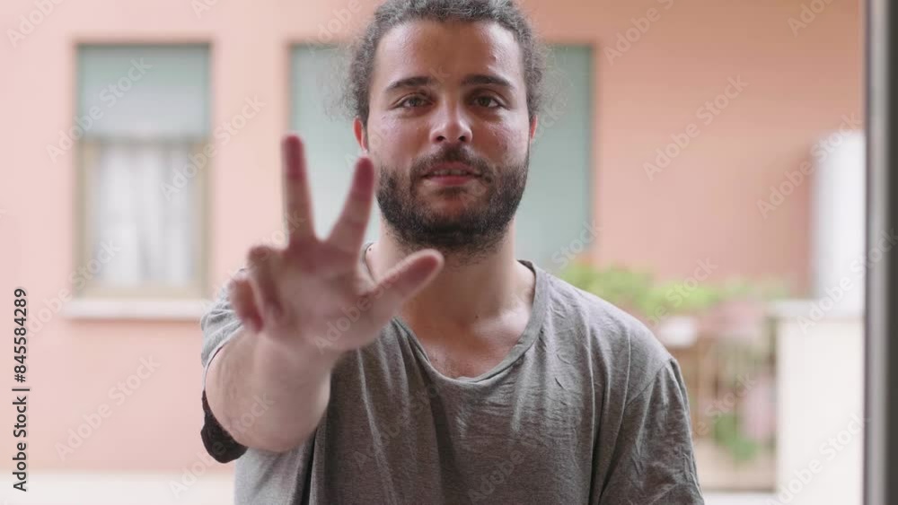Man in his 30s with long hair tied back and a scruffy beard, wearing a gray shirt, using his hands to count down from 5 to 0. He is standing on his home terrace with buildings in the background. Man in his 30s with long hair tied back and a scruffy beard, wearing a gray shirt, using his hands to count down from 5 to 0. He is standing on his home terrace with buildings in the background.