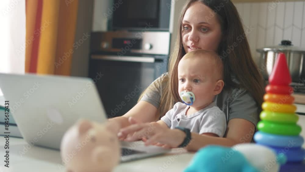 mother with a child sits at a table and works on a laptop. remote work mom baby concept. a woman works in a family setting lifestyle on the table are her child's toys. computer work mother with a child sits at a table and works on a laptop. remote work mom baby concept. a woman works in a family setting lifestyle on the table are her child's toys. computer work