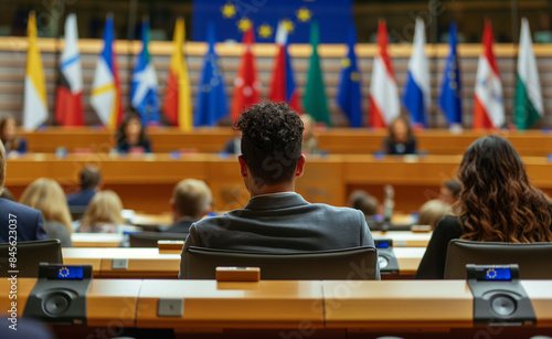 A man in a formal suit sits in a European parliamentary assembly, observing the proceedings.