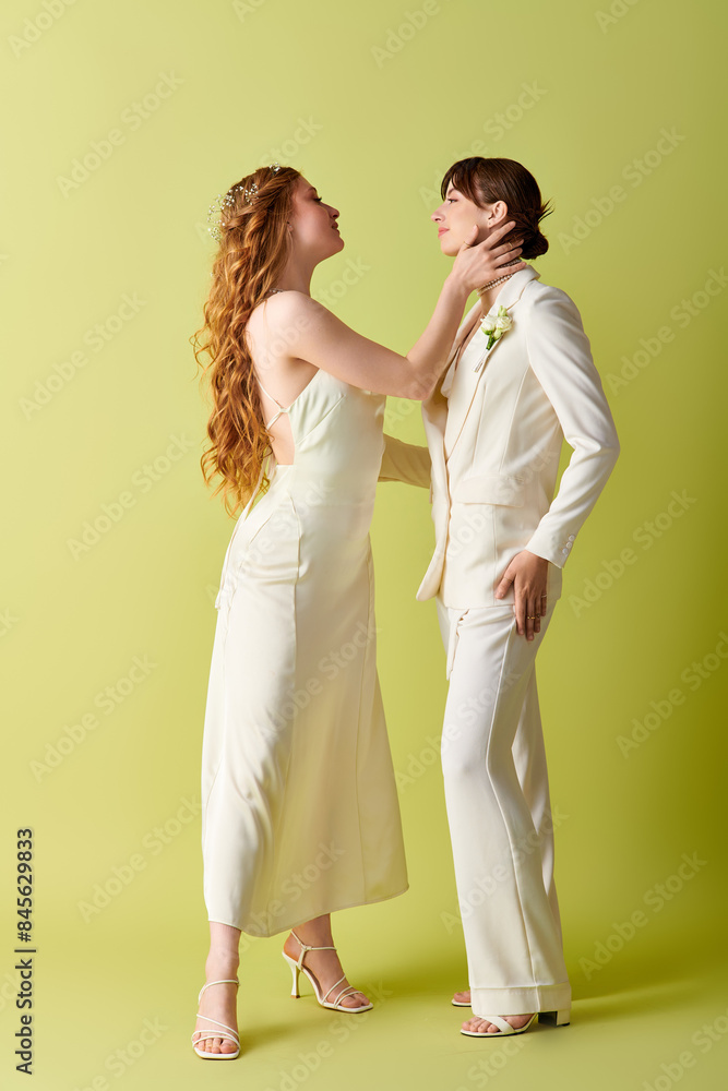 A lesbian couple dressed in white wedding attire stand facing each other against a green backdrop. A lesbian couple dressed in white wedding attire stand facing each other against a green backdrop.