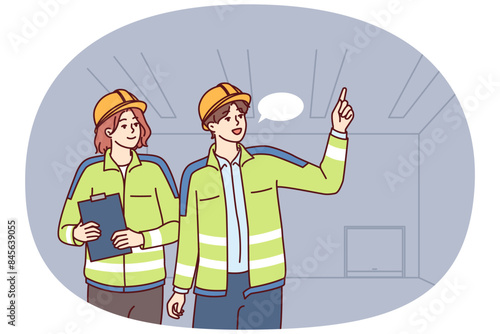 Two industrial room engineers discussing production process and how to repair equipment. Man and woman engineers in construction uniforms and safety helmets hold meetings in unfinished building