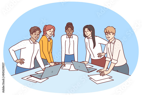 Team diverse business people stand at table with papers and laptops discussing company marketing strategy. Meeting in modern startup with team of young men and women engaged in corporate consulting