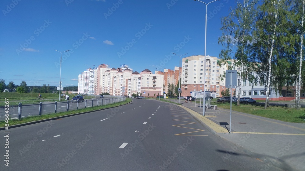 Along the city street there are residential houses, transportation stop and trees. Nearby paved sidewalk, bike lane and driveway with parked cars, lampposts. Sunny summer weather and blue sky Along the city street there are residential houses, transportation stop and trees. Nearby paved sidewalk, bike lane and driveway with parked cars, lampposts. Sunny summer weather and blue sky