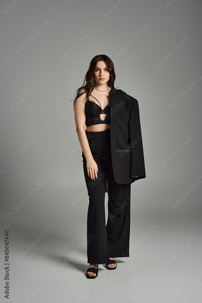 A stunning plus size woman strikes a pose in a sleek black top and pants against a gray backdrop. A stunning plus size woman strikes a pose in a sleek black top and pants against a gray backdrop.