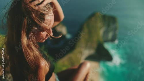 A woman with long hair is sitting on a cliff overlooking the ocean, filmed in slow motion at Kelingking Secret Point Beach, Nusa Penida Island, Indonesia. Female tourist on Bali summer vacation. photo