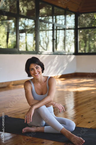 Young happy fit sporty slim Hispanic woman yoga coach, stretching sport trainer wearing grey top and pants sitting on mat in modern yoga gym studio in nature park looking at camera. Vertical portrait. photo