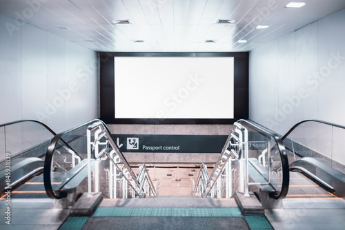 Two escalators and staircase leading down to a passport control area with a large blank billboard above for advertisement mockup. The scene is set in a modern, well-lit airport terminal interior